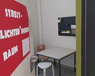 Ein kleiner Raum mit einem weißen Tisch, zwei Stühlen und einer Pinnwand an der Wand