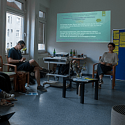 Fünf Personen sitzen in einem Raum und sehen eine Wandpräsentation mit Text und Bildern