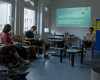 Fünf Personen sitzen in einem Raum, während eine Präsentation an die Wand projiziert wird